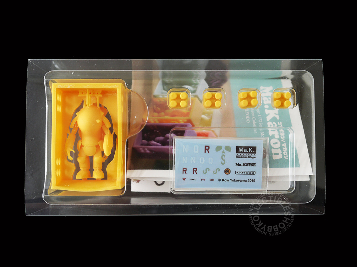 A packaged 3D-printed Kaiyodo Ma.K 1/72 scale Ma.Karon version SAFS kit Fire Ball SG Orange color figure set in clear plastic blister packaging on a black background. The set includes a small Orange SAFS figure, four hexagonal studs, printed instruction manual and decals sheet.