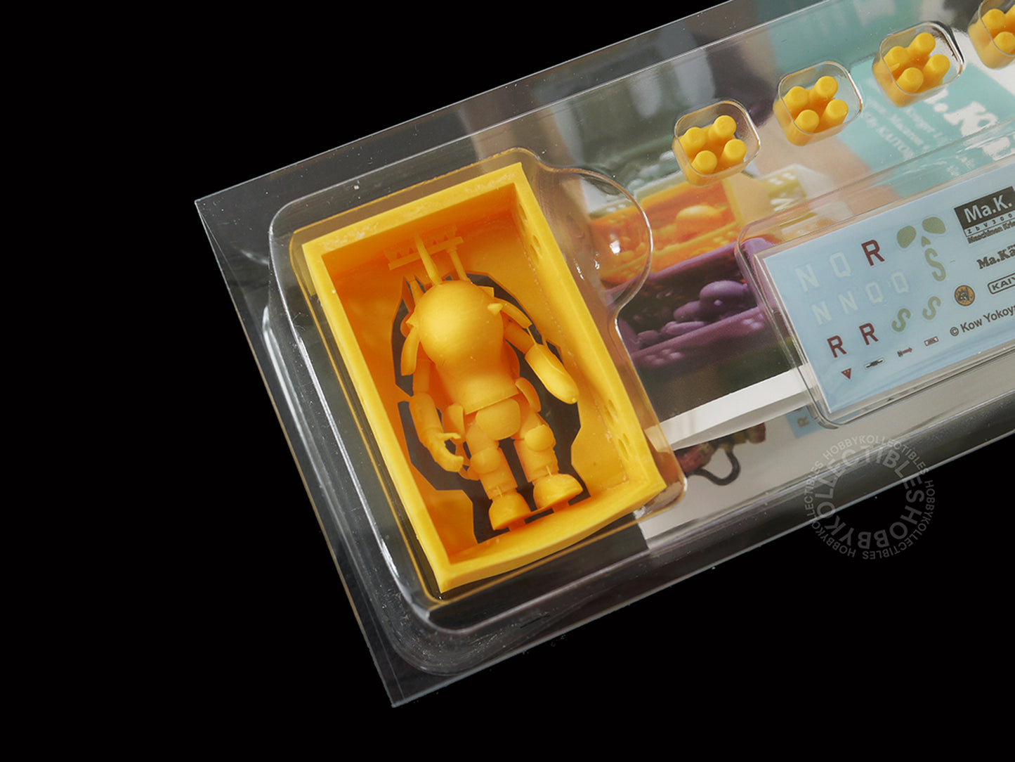 Close up shot of  3D-printed Kaiyodo Ma.K 1/72 scale Ma.Karon version SAFS kit Fire Ball SG Orange color figure set in clear plastic blister packaging on a black background. The set includes a small Orange SAFS figure, four hexagonal studs, printed instruction manual and decals sheet.